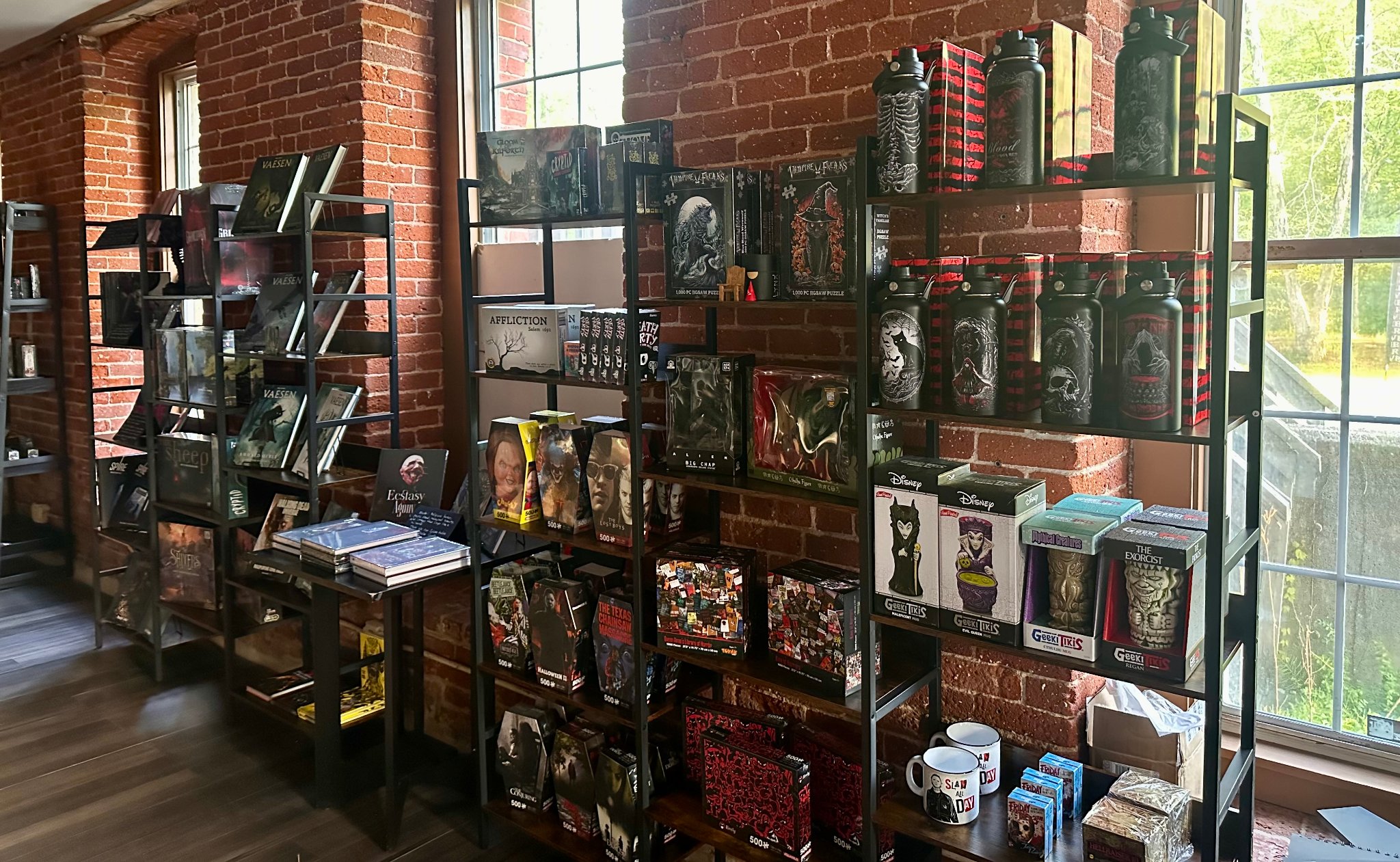 Wide view of The Cursed Page merchandise wall — horror-themed puzzles, collectibles, and gothic water bottles on black shelves against exposed brick