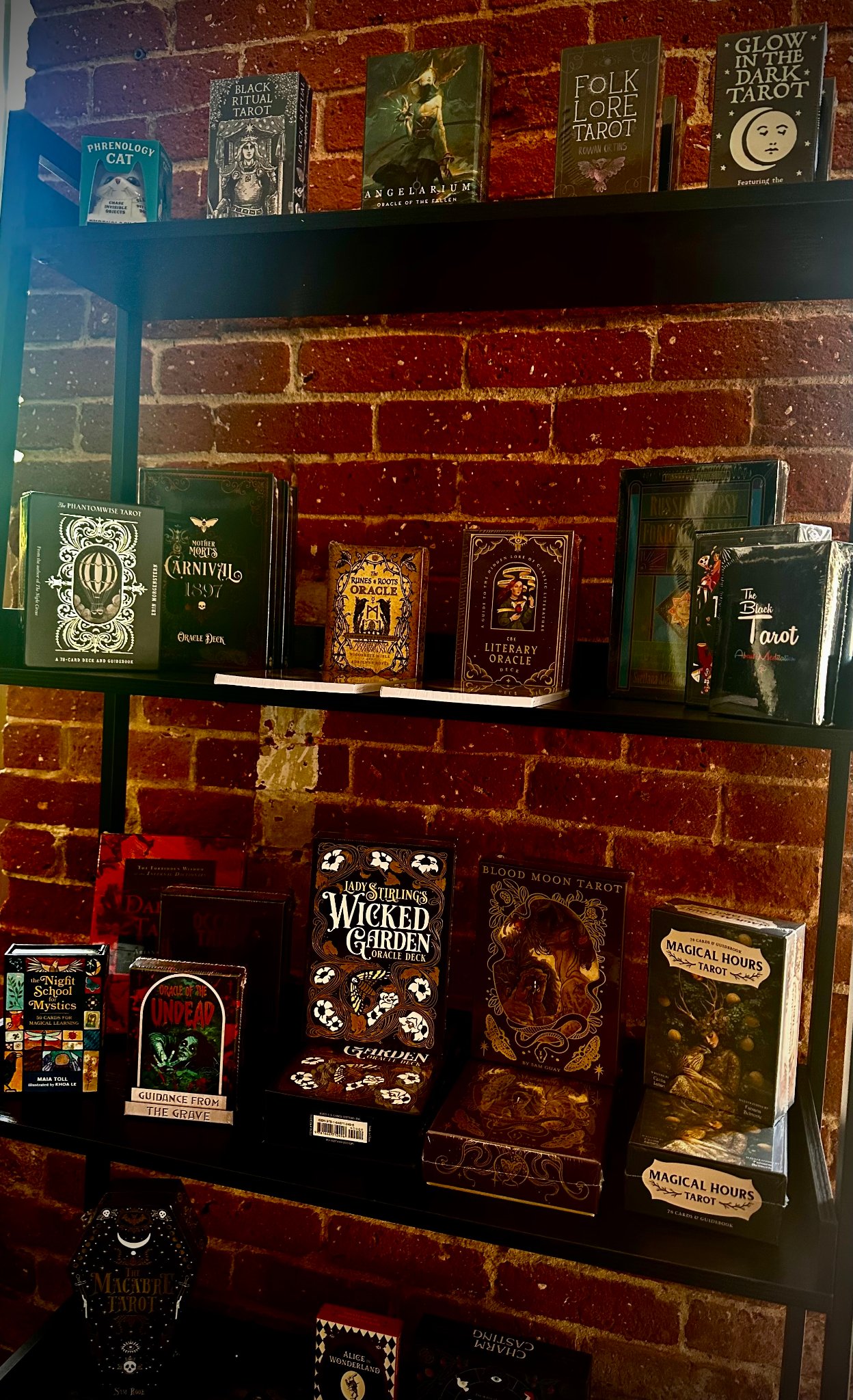 Tarot and oracle card shelf — Black Ritual Tarot, Folklore Tarot, Literary Oracle, Blood Moon Tarot, and more against exposed brick