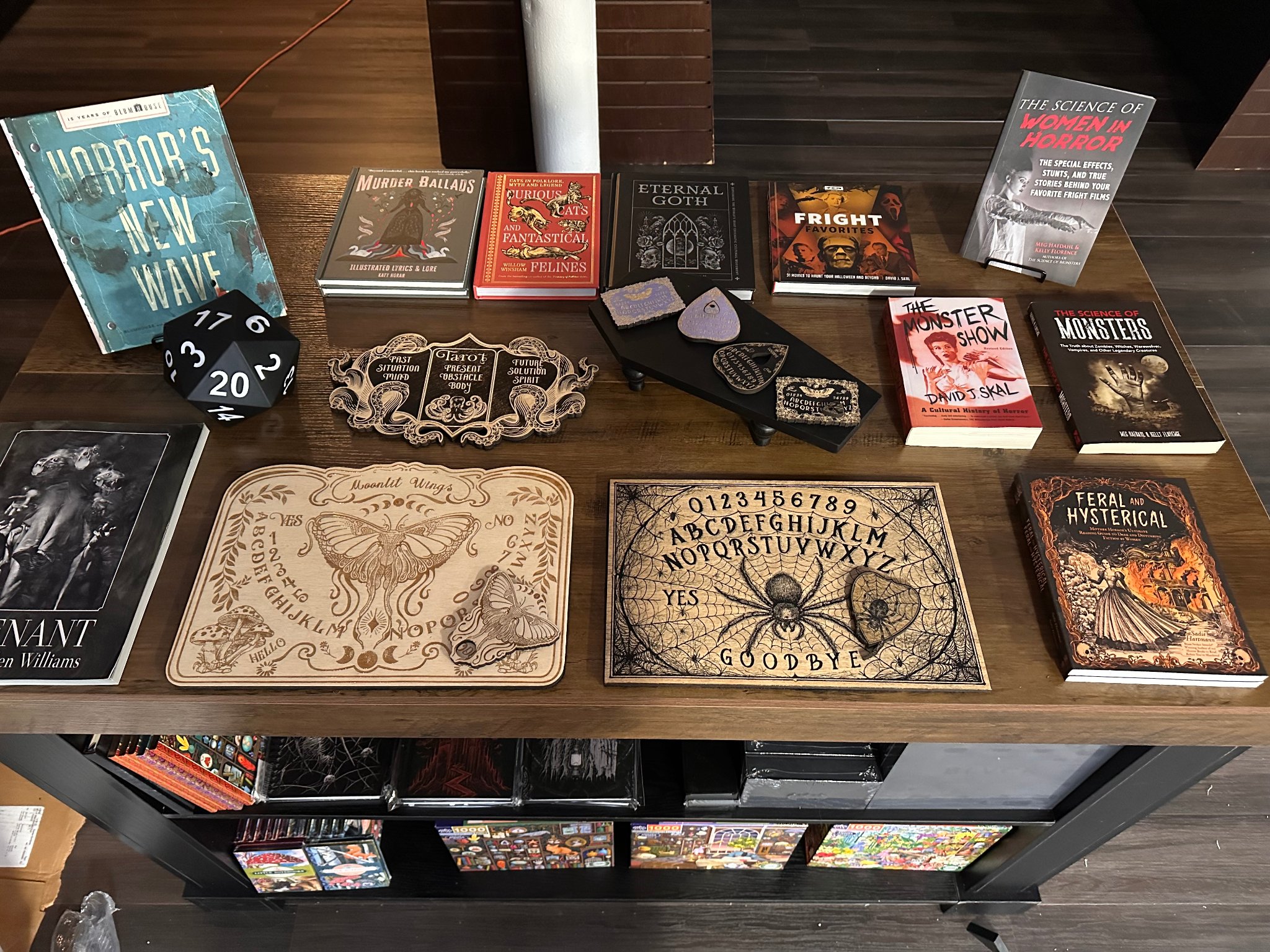 Merchandise table display featuring Ouija boards, horror books, a d20 die, and gothic curiosities